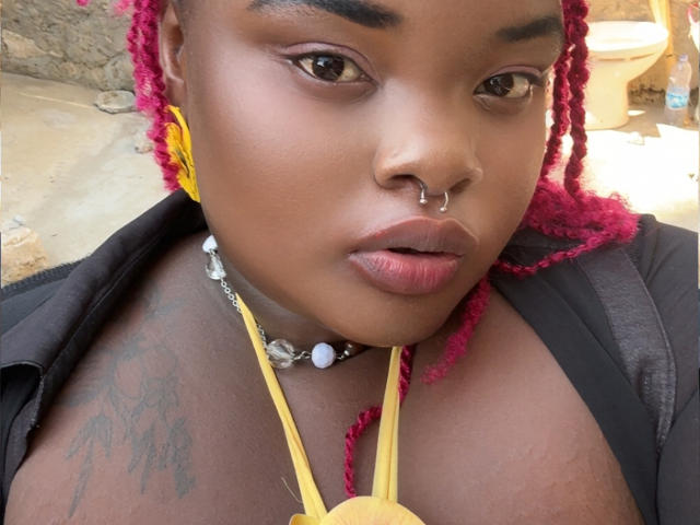 Close-up photo of BigChocoTitiey, a black woman with red dreadlocks, nose ring, and dark lipstick, wearing a black shirt, yellow necklace, and beaded choker, with a visible tattoo on her chest. Fucking intense gaze.
