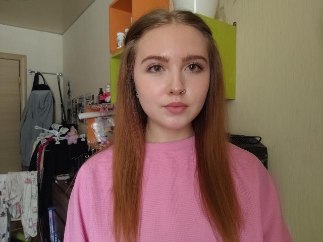 TerraMaul with long brown hair, wearing a pink sweater, has a nose ring, and a slightly flushed face. Background: cluttered room with clothes and shelves. Fucking cute, sexy, innocent look.