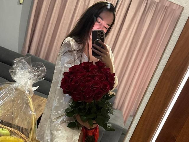 AraiaSheri prend un selfie devant un miroir, tenant un grand bouquet de roses rouge foncé. Elle porte un haut blanc transparent, avec de longs cheveux noirs et une expression séduisante, son téléphone couvrant partiellement son visage..