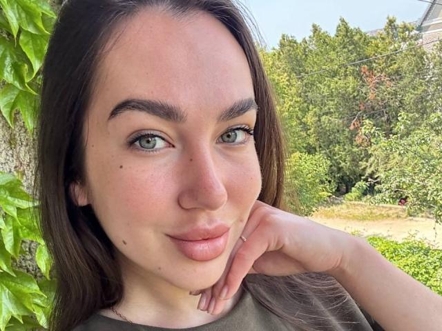 Close-up of AraiaSheri, a fair-skinned woman with green eyes, dark brown hair, and a slight smile. She rests her hand on her cheek, showing a few moles. Outdoor greenery background. Fucking gorgeous.