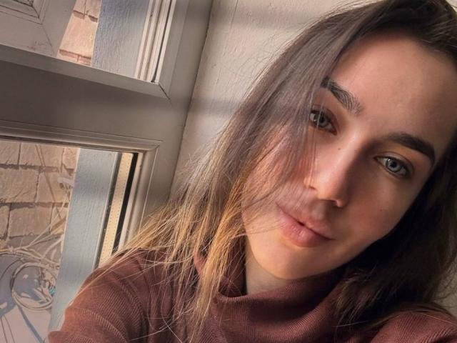 Close-up of AraiaSheri, a young woman with long brown hair, light skin, and blue eyes, leaning against a window frame. Sunlight casts shadows across her face, highlighting her relaxed, slightly seductive expression.