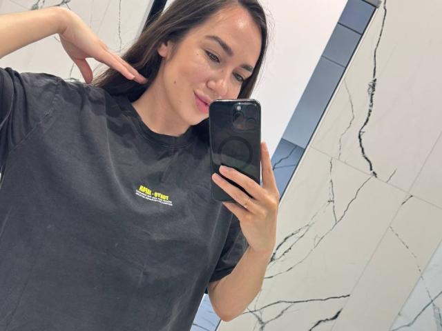 AraiaSheri, con i suoi lunghi capelli castani, si scatta un selfie in una stanza ben illuminata. Indossa una maglietta nera, ha una piccola lentiggine sulla guancia e si tocca l'orecchio con una mano. Il suo telefono è nero.
.