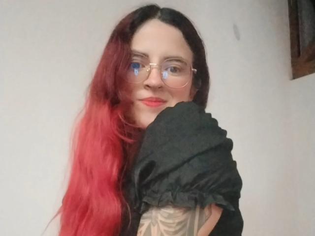 Photograph of LaiaJonnes, pale-skinned with red and black hair, wearing glasses and black top, exposing tattooed arm, leaning forward, smiling slightly, creating sexual tension, casual pose, soft lighting.