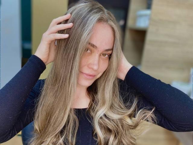 Melanie, avec ses longs cheveux blonds ondulés, porte un haut noir à manches longues, les mains dans les cheveux, et regarde directement la caméra, léger sourire, lumière douce, intérieur, tension sexuelle.