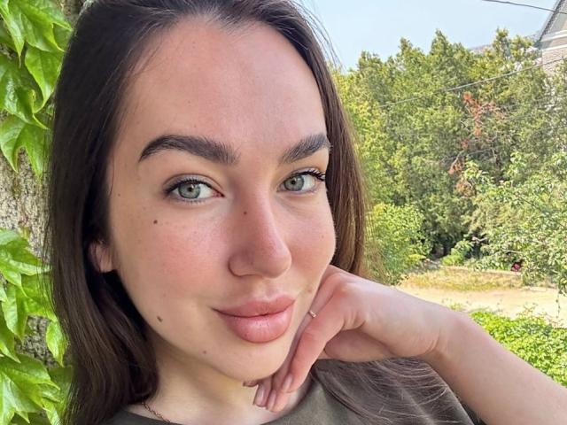 Close-up van AraiaSheri, een jonge vrouw met groene ogen, donkerbruin haar en lichte huid, die leunt tegen een groene muur. Hand rustend op haar wang, groene planten op de achtergrond..