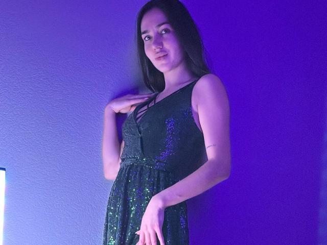 AraiaSheri steht vor einer blau beleuchteten Wand und trägt ein schwarzes Kleid mit Pailletten. Sie ist leicht gedreht, die Hand auf der Brust, und strahlt eine sexuelle Spannung aus. Ihre dunklen Haare bilden einen Kontrast zum leuchtenden blauen Licht..