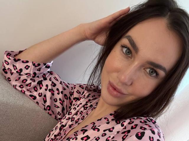 Close-up of AraiaSheri, a white woman with green eyes, dark brown hair, and a light pink leopard print top. She's leaning against a gray couch, hand in hair, sexy smile.