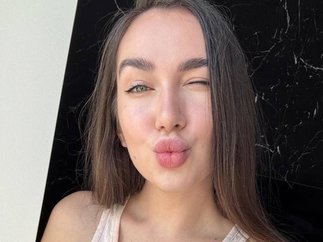 Close-up of AraiaSheri, a young woman with long dark hair, winking and pouting her lips. She's wearing a white tank top, against a black cracked background. Fucking cute, sexy face.