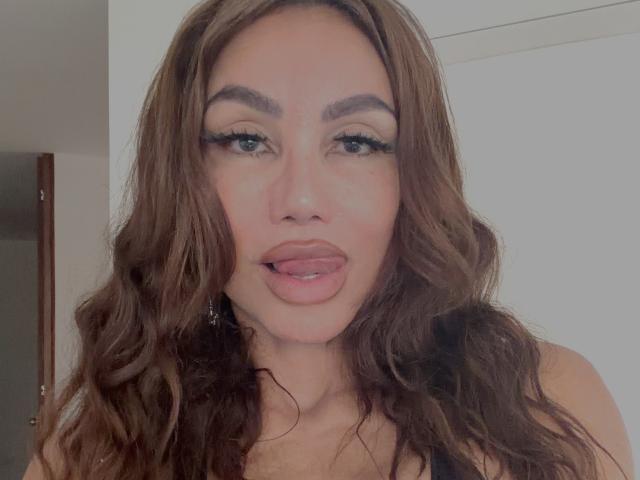 Gros plan de Paulatina, avec de longs cheveux bruns ondulés, des lèvres pulpeuses légèrement entrouvertes et un trait d'eye-liner prononcé. Elle regarde directement l'appareil photo avec une expression séduisante. Éclairage vif, fond neutre
.