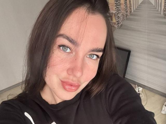 Close-up selfie of AraiaSheri, a white woman with long black hair, blue eyes, and full pink lips. She wears a black hoodie, looking directly at the camera with a slight smirk. Background includes wooden stairs. Sexual tension from direct gaze and slight smirk.