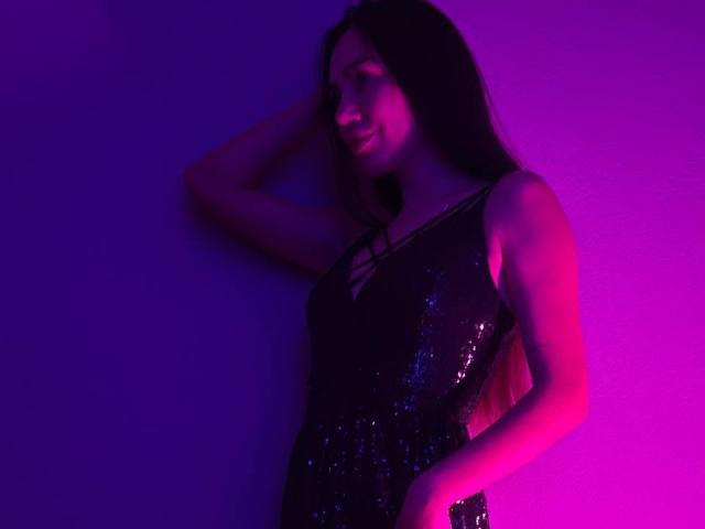 AraiaSheri, mit ihren langen, dunklen Haaren, lehnt an einer Wand und trägt ein schwarzes, pailletenbesetztes Kleid, das im violetten und rosa Neonlicht erstrahlt. Licht.
ihr Arm ist erhoben, ein Ausschnitt des BH ist unter dem Kleid sichtbar..