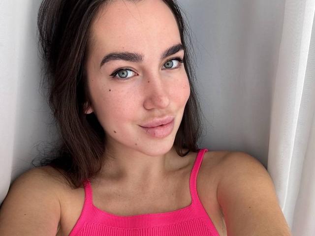 Gros plan d'AraiaSheri, une jeune femme aux longs cheveux noirs, portant un débardeur rose vif, à la peau claire et aux taches de rousseur. Elle a un sourire subtil, des yeux verts et une petite tache de naissance visible sur sa joue..