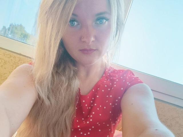 Photograph of NicoleDreamss with long, blonde hair, wearing a red polka dot shirt, sunlight illuminating her face, blue eyes, light skin, close-up selfie, sexual tension, natural light, window in background.