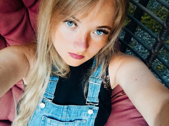 Nahaufnahme von NicoleDreamss, einer blonden Frau mit blauen Augen, die ein schwarzes Oberteil und blaue Jeans-Overalls trägt und auf einer rotes Kissen, das in die Kamera blickt und einen leicht verführerischen Ausdruck hat..