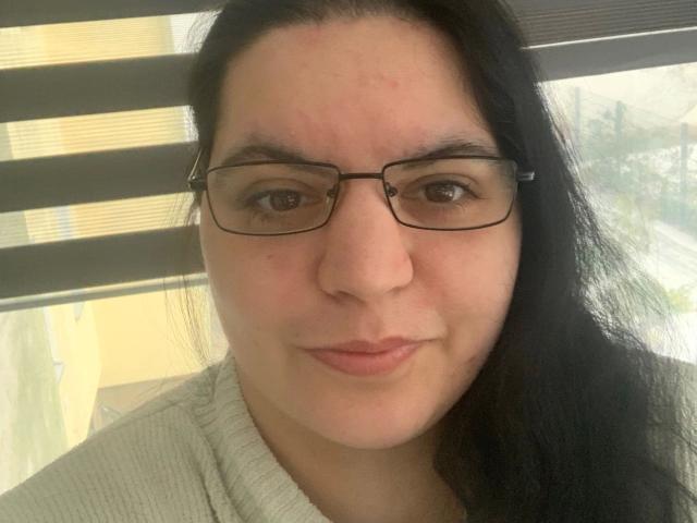 Close-up selfie of Curvy-jasmin1990-hot, a Latina woman with long black hair, wearing glasses and a light gray sweater. She has a slight smile and visible pores on her face. Background includes striped blinds.