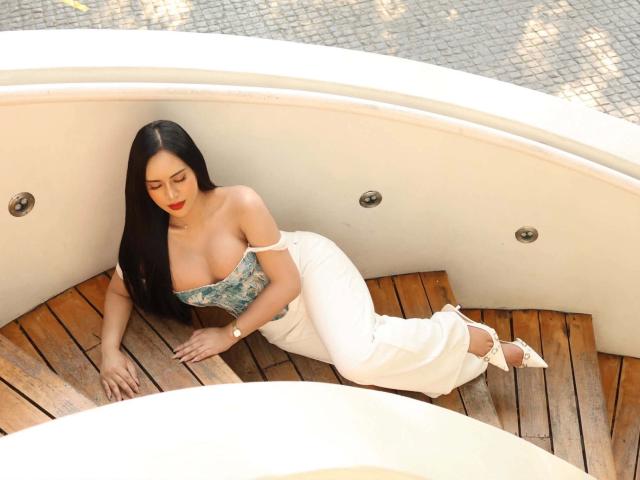Madison Cassanova, assise sur un banc en bois dans un bateau, est vêtue d'un haut bleu sans bretelles avec un décolleté plongeant, et d'un pantalon blanc. La lumière du soleil met en valeur ses longs cheveux noirs et son rouge à lèvres. Incroyablement sexy..