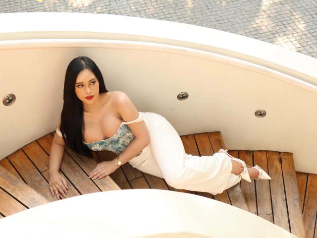 Madison Cassanova, con un aspecto atractivo, está recostada sobre una cubierta de madera curvada, vistiendo una blusa verde sin tirantes que deja al descubierto sus grandes senos y su piel blanca. pantalones. La luz del sol resalta su cabello oscuro y sus labios rojos, creando una vibra sexy y relajada..
