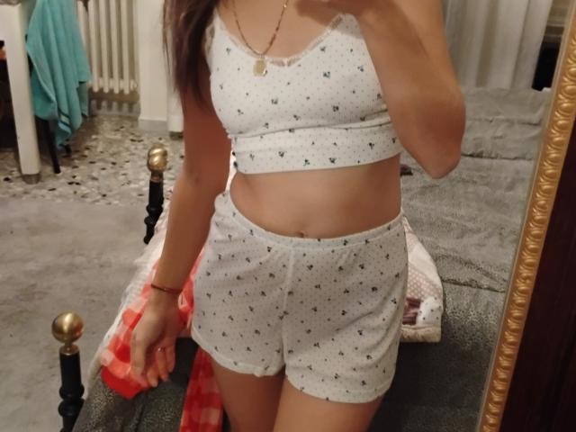 AliiceX maakt een selfie voor een spiegel, gekleed in een witte top met een hoge hals en bijpassende korte broek met kleine zwarte patronen. Ze houdt een rood-wit geruit stuk stof vast, waardoor haar platte buik en slanke figuur zichtbaar worden..