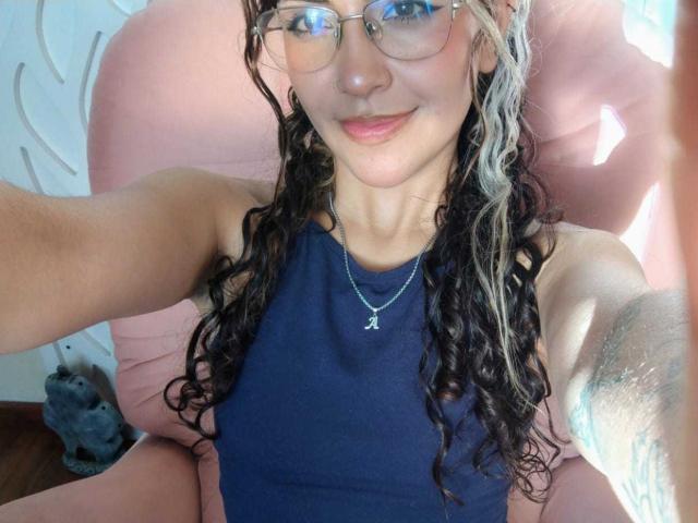 AdaraAngel, una donna esile con lunghi capelli ricci castani, che indossa occhiali e una canotta nera, è seduta su una sedia rosa. Sorride, e indossa una collana d'argento con un ciondolo a forma di croce. La luce del sole illumina il suo viso.