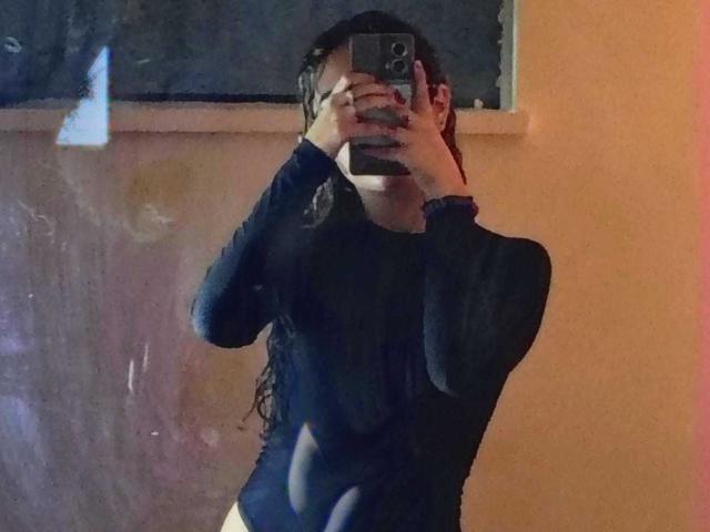 AdaraAngel, eine schlanke Person mit langen, feuchten, lockigen Haaren, trägt ein eng anliegendes, schwarzes Oberteil mit langen Ärmeln und macht ein Selfie mit dem Spiegel. mit einer Hand, die ihr Gesicht verdeckt, wobei ein schwach beleuchteter Raum reflektiert wird.