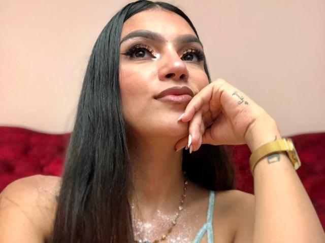 Close-up of NoemiFerrer, a woman with long black hair, sparkling skin, and a light blue top, resting her chin on her hand, wearing a gold wristwatch, looking upwards with a seductive expression.