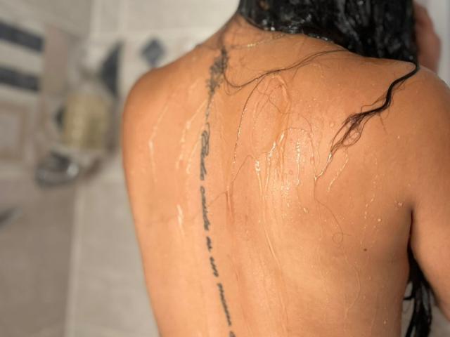 Close-up das costas de RouxiMori, molhadas e brilhantes, com uma linha de tatuagem preta que percorre o centro. Cabelo escuro e úmido cai para a direita. O tom de pele é bronzeado. Há uma leve tensão erótica..