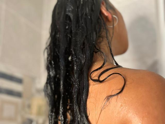 Primer plano del cabello negro y mojado de RouxiMori, su hombro y su oreja, con gotas de agua visibles. El fondo está desenfocado, centrándose en la textura de la piel y el cabello húmedos. Tensión sugestiva, erótica..