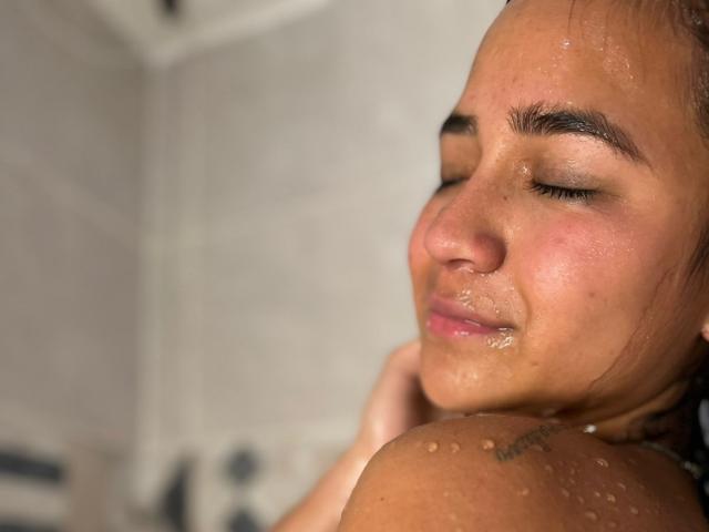 Photo en gros plan de RouxiMori, une femme attirante, transpirante, les yeux fermés et un léger sourire, montrant une peau humide et petit tatouage sur son épaule. Concentrez-vous sur son expression détendue et séduisante..