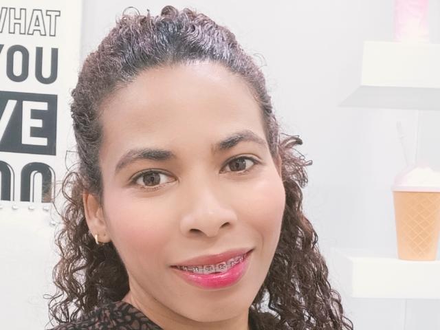 Close-up foto van RebecaDuval, een mollige vrouw met krullend donker haar, medium bruine huid en roze lippenstift, die lacht. direct in de camera, met een witte achtergrond en vetgedrukte zwarte tekst.