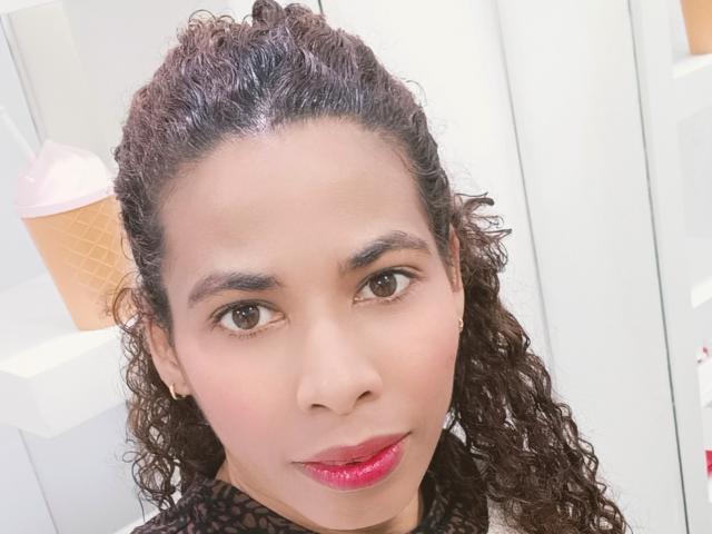 Close-up of RebecaDuval, a curvy woman with curly dark hair, olive skin, and red lipstick. She's looking directly at the camera with a slight smile, her dark eyes intense. Bright background.