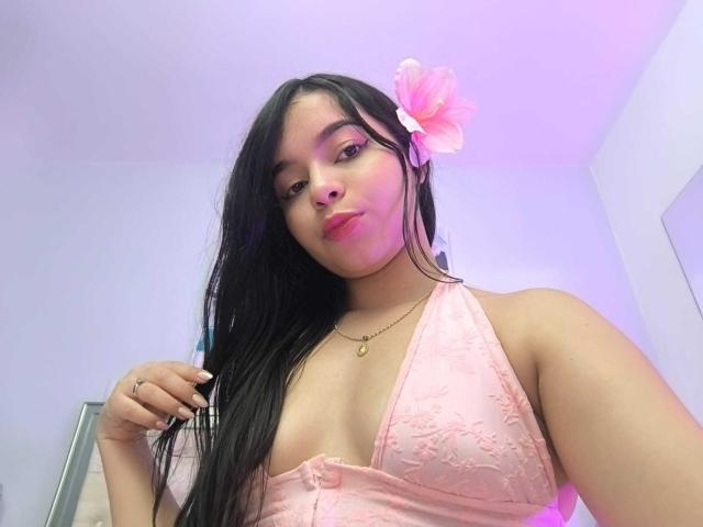 Lissy Carter mit langem, schwarzem Haar, trägt ein pinkes Spitzen-Haltertop, eine pinke Blume im Haar und blickt subtil in die Kamera. Lächeln, sichtbarer Ausschnitt, weiches Licht, leichtes rosa Schimmern im Hintergrund..