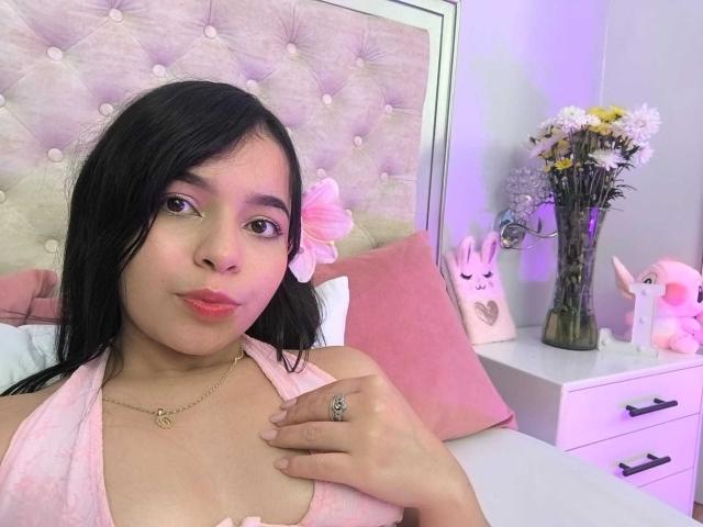 Lissy Carter, com cabelo preto, pele clara e batom vermelho, vestindo uma blusa rosa de alças, com a mão no peito, um anel visível, cabeceira com detalhes em relevo, almofadas rosa, vaso com flores, decoração com coelhos, iluminação suave.
