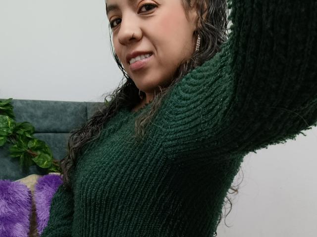 Autoportrait en gros plan de MeganFerrerr, avec de longs cheveux bruns bouclés, portant un pull vert en tricot, levant son bras droit. montrant un piercing au nez, souriant, avec un canapé gris et une plante verte en arrière-plan..