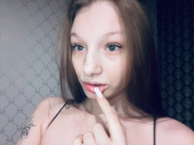 Nahaufnahme von MelbaCaffee, einer jungen Frau mit heller Haut und braunen Haaren, die mit ihrem Finger eine Zahnbürste ableckt, wobei der Finger im Mund ist. Sie trägt ein schwarzes Top mit Trägern, und im Hintergrund ist eine Wand mit einem Muster zu sehen..