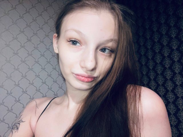 Close-up of MelbaCaffee, a fair-skinned woman with long brown hair, blue eyes, and pink lips, wearing a black spaghetti strap top, slight smile, light tattoo on shoulder, soft shadowing.