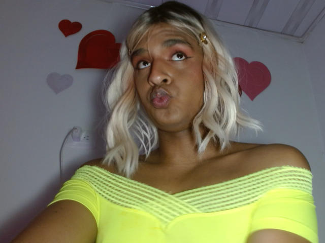 Photograph of IdiiaLatina with medium brown skin, pouty lips, and platinum blonde hair. Wearing a neon yellow off-shoulder top. Red and purple heart stickers on the wall. Fucking cute.