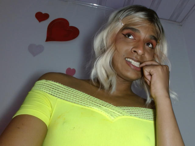 Photograph of IdiiaLatina, a young woman with light brown skin and platinum blonde hair, wearing a bright yellow off-shoulder top, smiling seductively, biting her finger, red and pink hearts in background.