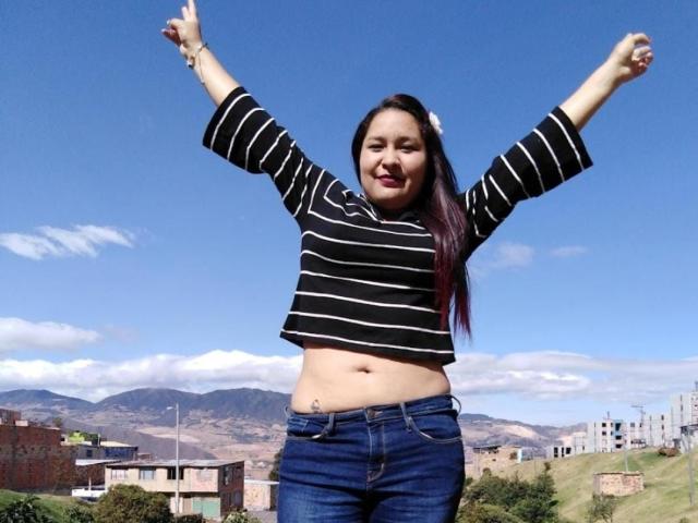 CattaleyaBohorquez stands with arms raised, smiling, wearing a black and white striped crop top and blue jeans. Clear blue sky, distant mountains, and buildings in the background. Bright sunlight.