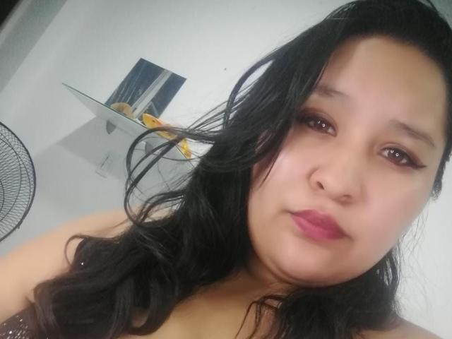 Close-up selfie of CattaleyaBohorquez, Asian woman with long black hair, pink lipstick, and brown eyes. She has a slightly pouty expression, wearing a black top, with a fan and yellow fruits in the background.
