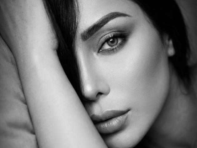 Nahaufnahme von SandraXSeins, ein Schwarz-Weiß-Foto. Sie hat die Hälfte ihres Gesichts im Schatten, einen intensiven Blick und leicht geöffnete Lippen. Ihre Haut ist glatt, ihre Augen stark geschminkt. Der Unterarm bedeckt einen Teil ihres Gesichts. Sexuelle Spannung.