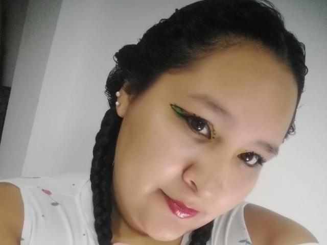 Close-up selfie of CattaleyaBohorquez, an Asian woman with black braids, green eyeshadow, and red lipstick, smiling with a hint of sexual tension, wearing a white top. Bright lighting.