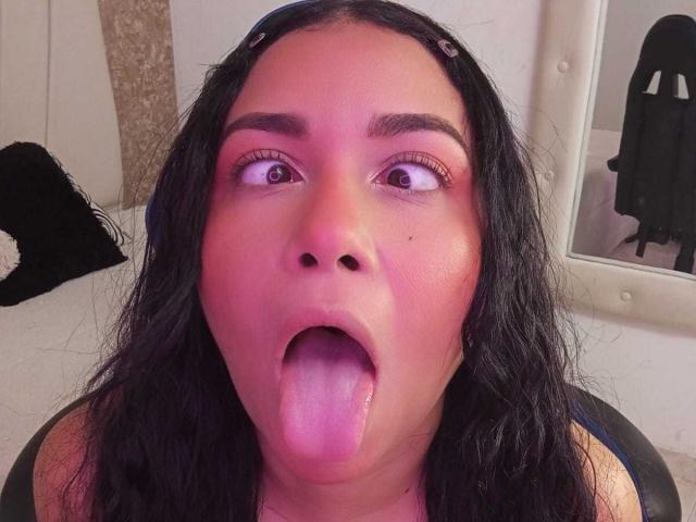 Foto em close-up de Alice Smity, com cabelo escuro, olhos grandes, língua de fora, pele rosada-avermelhada, roupa preta e espelho ao fundo. Sua expressão é sexual e provocativa..