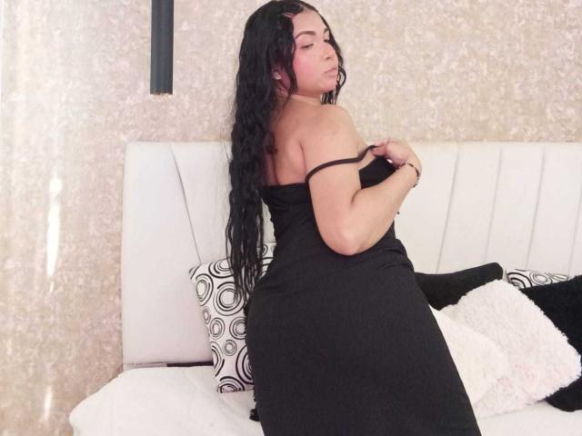 Alice Smity, uma mulher curvilínea com longos cabelos pretos, vestindo um vestido preto sem alças, sentada em um sofá branco, ligeiramente virada, puxando a alça do vestido, expressão confiante, sexy, iluminação suave, decoração minimalista..