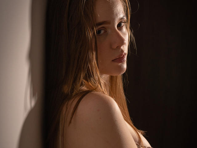 Foto van ArielLoveX met nat bruin haar, in profiel, met een intense blik in de camera. Het licht accentueert haar rechter schouder en wang, en werpt schaduwen. Donkere achtergrond. Seksuele spanning, verleidelijke uitdrukking..