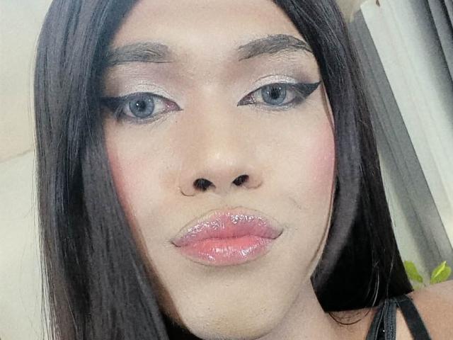 Close-up van UnforgetableAnna met glanzende, roze lippen, dikke zwarte eyeliner en glinsterende oogschaduw. Ze heeft lang zwart haar, bleke huid en blauwe ogen, en kijkt recht in de camera met een verleidelijke uitdrukking..