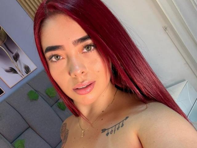 Autoritratto ravvicinato di ValeriaRoseT con lunghi capelli rossi, pelle chiara e tatuaggi sul petto. È senza top, con una piccola collana d'oro. La sua espressione è leggermente seducente. Luce interna intensa.