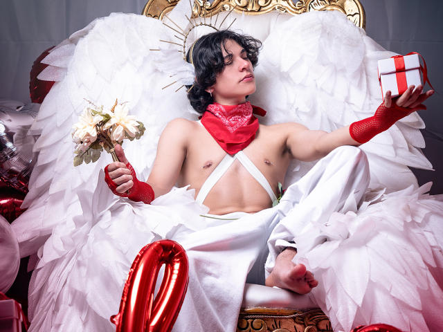 Luke Howlandy, musclé et torse nu, porte un costume blanc orné de plumes et d'accessoires rouges, et tient une boîte cadeau. Des ballons rouges et des touches dorées. Assise sur une chaise ornée, elle a l'air confiante et séduisante..