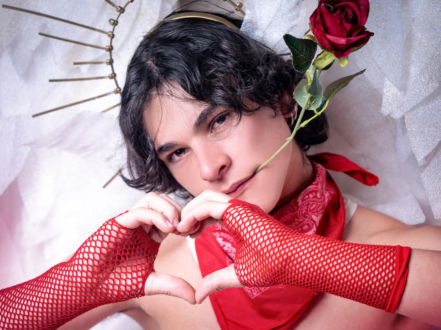 LukeHowlandy, liggend op een wit bed, draagt rode handschoenen van netstof en een rode sjaal, houdt een rode roos vast, heeft zwart krullend haar en een kroon. met gouden spikes, suggestieve uitdrukking, direct oogcontact, intense seksuele spanning.