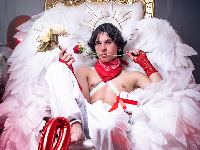 Luke Howlandy, com cabelo preto, está sentado em uma cadeira ornamentada, vestindo uma fantasia branca com penas, um lenço vermelho e meia-calça de rede. luvas, revelando um falo, cercado por fitas vermelhas e penas brancas.