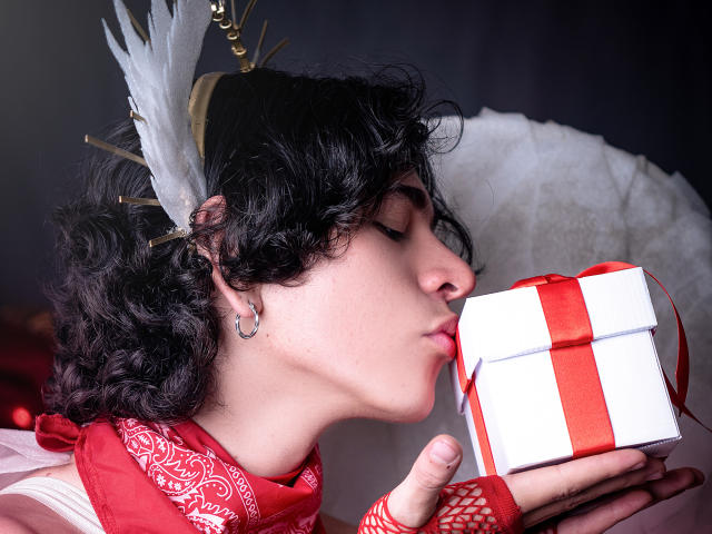 Luke Howlandy, con cabello negro rizado y un tocado de plumas blancas, besa un regalo envuelto en papel rojo y blanco, vistiendo encaje rojo. Guante y bufanda roja. Fondo oscuro, enfoque intenso en el regalo..